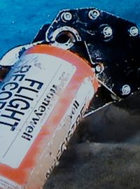 Mechanické rameno drží oranžový válcovitý záznamník letových parametrů nad pískem
